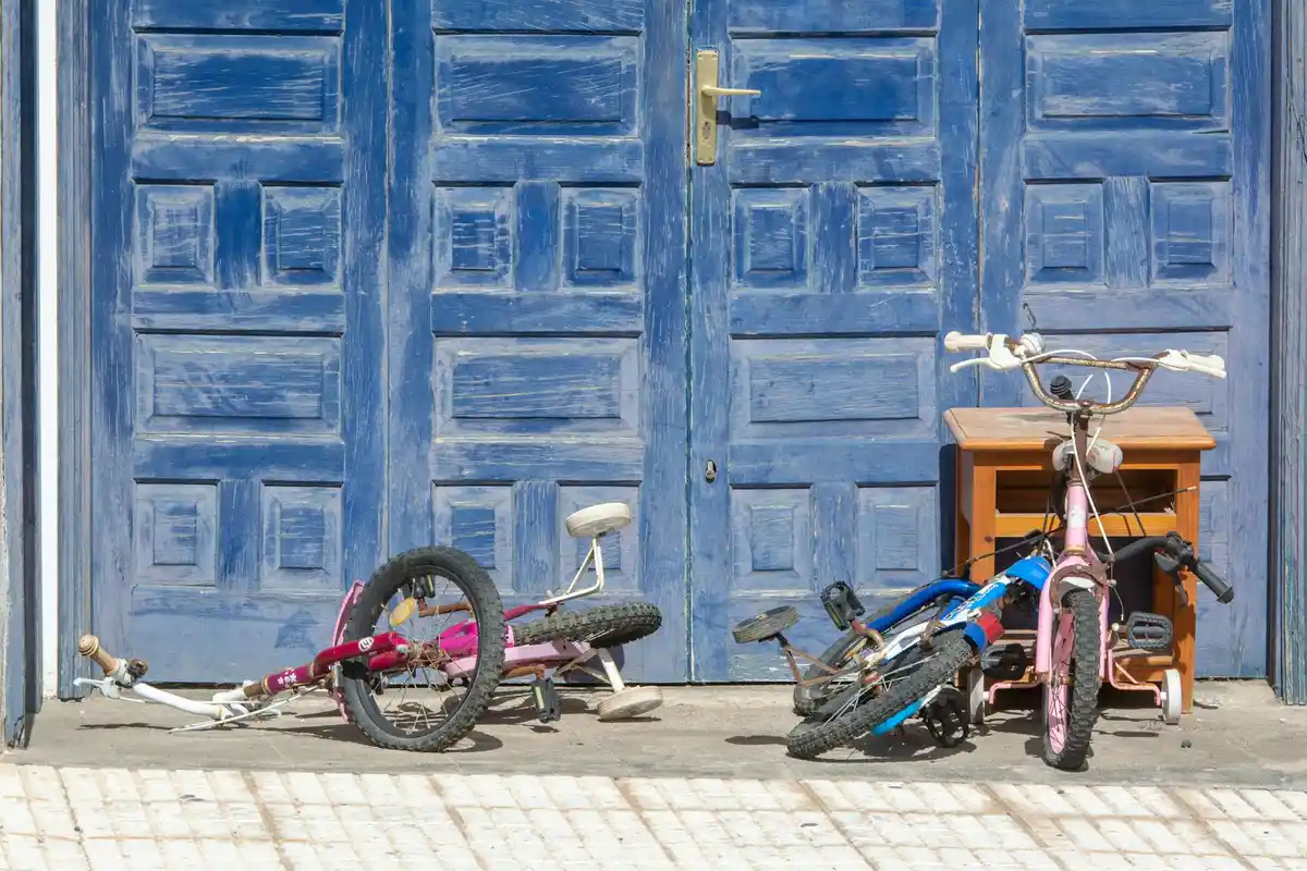 Two bicycles, one pink and one blue, lie on the ground in front of blue garage doors; a small wooden cabinet sits to the right.