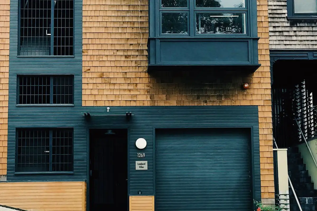 Blue garage door on a tan-shingled townhouse with a side entry door and stairs, typical residential garage setup.