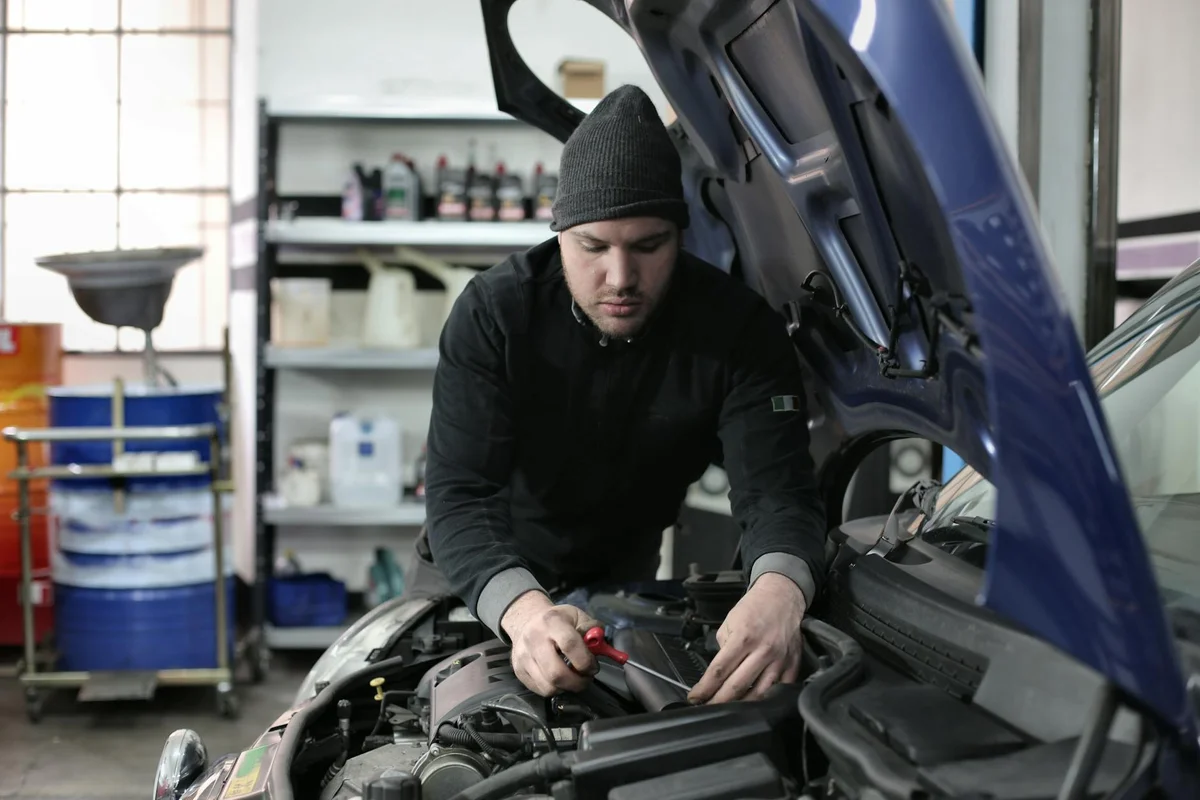 A mechanic wearing a beanie leans over the engine with the hood open in a workshop, inspecting the engine bay.