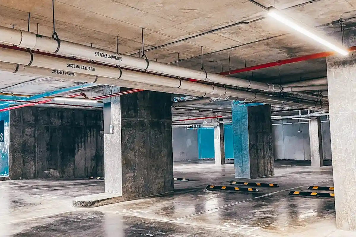 A vacant underground parking garage with exposed concrete columns, overhead pipes, and a glossy, damp-looking concrete floor.