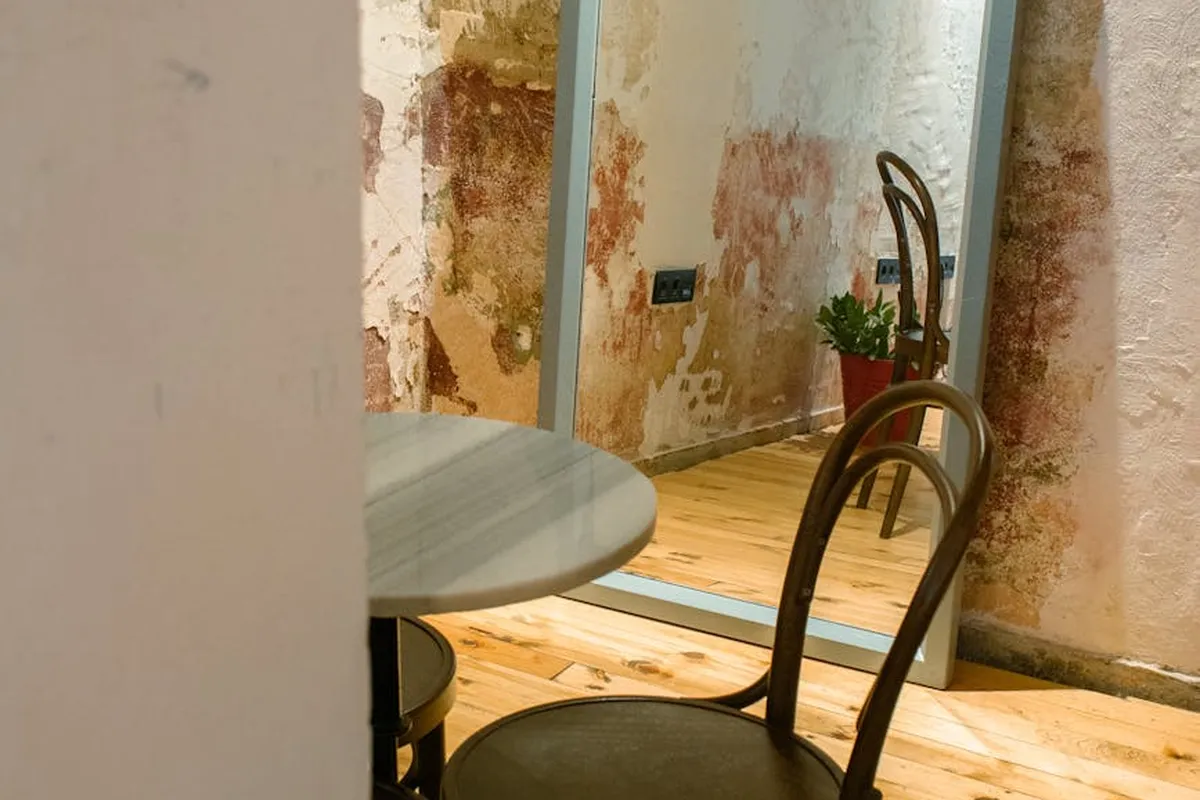 Interior of a small converted garage with a round table, two metal chairs, a large mirror, exposed brick walls, and wooden floors.