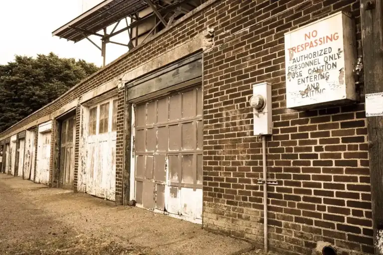 Exterior of a row of old detached brick garages with metal doors; a 'No Trespassing' sign is mounted on the wall.