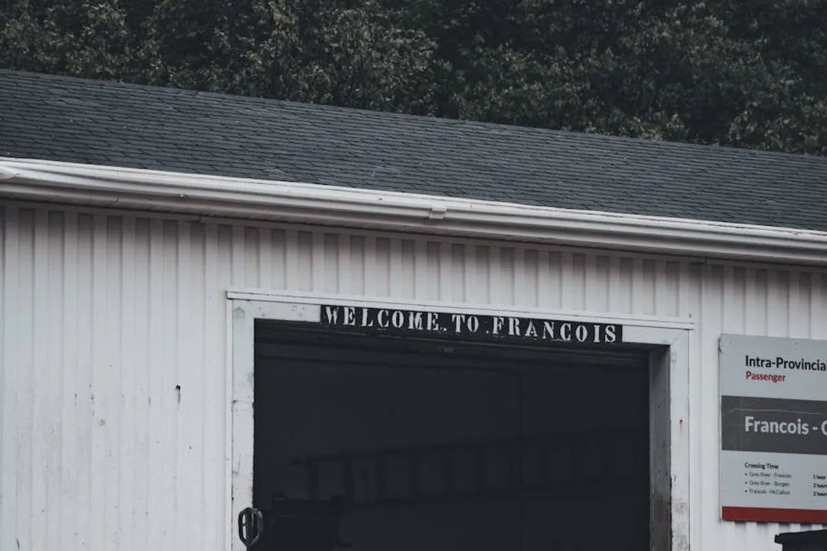 White metal garage building with an open doorway and a sign above reading 'WELCOME TO FRANCOIS'; a small information board is mounted to the right side of the entrance.