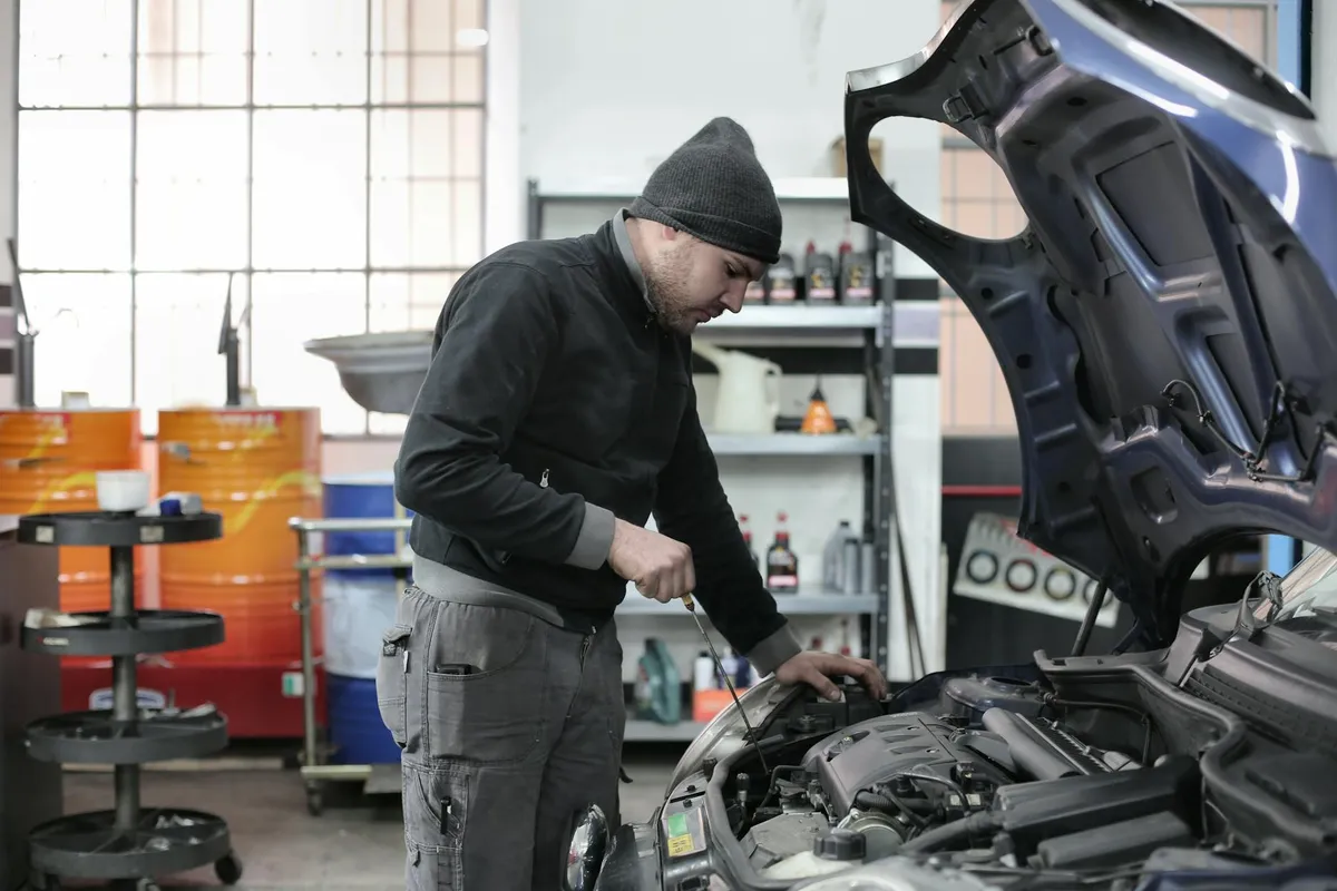 A mechanic wearing a beanie works on an open car hood inside a well-organized garage with shelves and tools in the background.