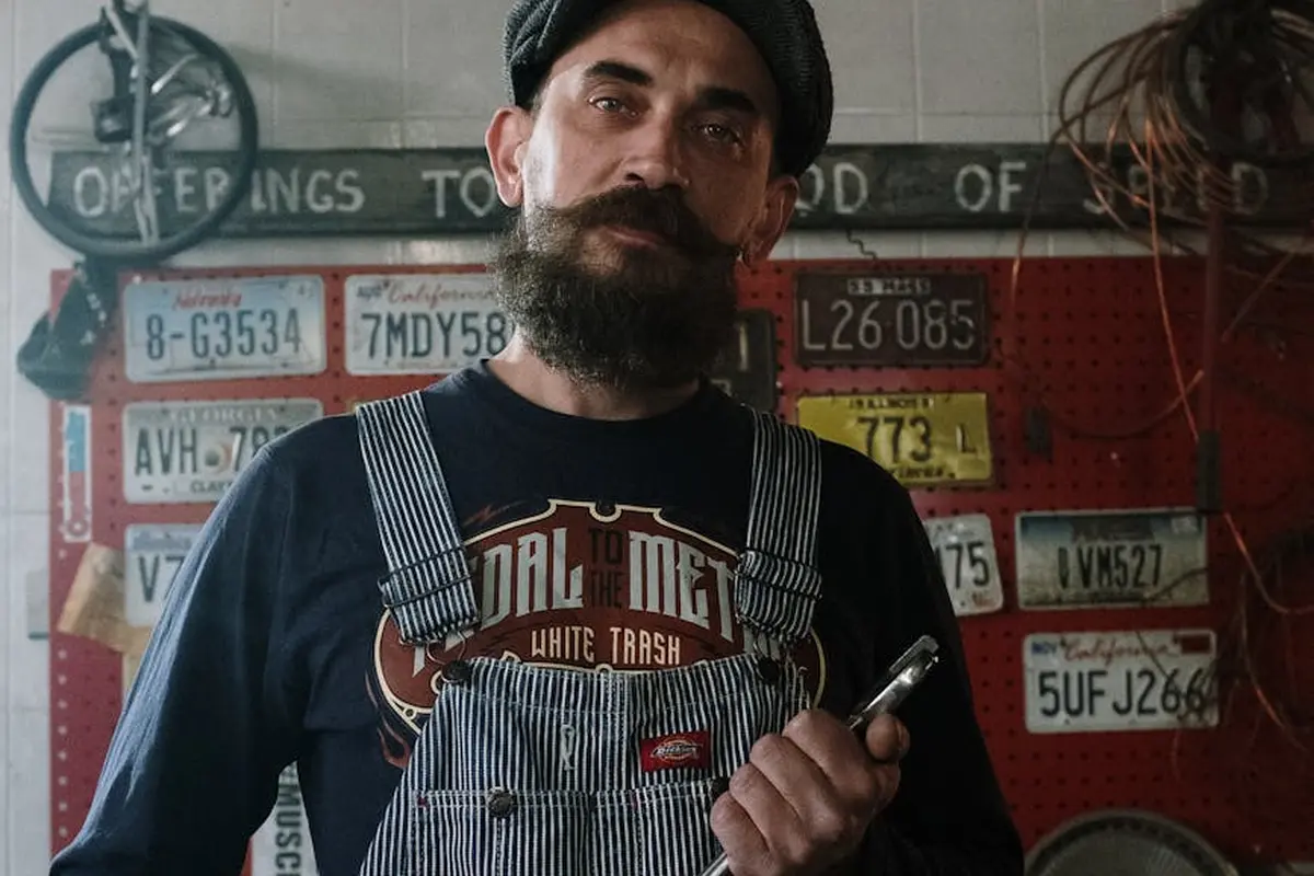 A bearded man wearing striped overalls and a dark T-shirt stands in a cluttered garage workshop, holding a tool, with license plates mounted on the wall behind him.