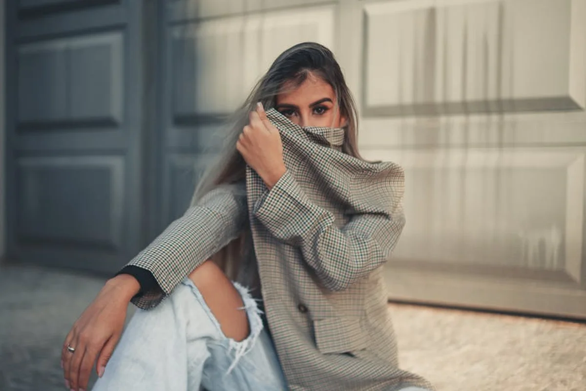 Person sitting on the ground in front of a garage door, wearing an oversized plaid blazer and ripped jeans, partially covering their face with their sleeve.