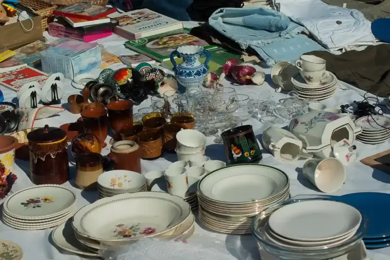 Assorted plates, bowls, mugs, teapots, and glassware scattered across a cluttered table in a garage.