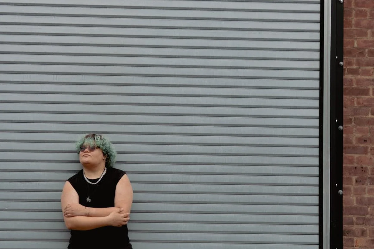 Person standing with arms crossed in front of a gray metal garage door, brick wall on the right.