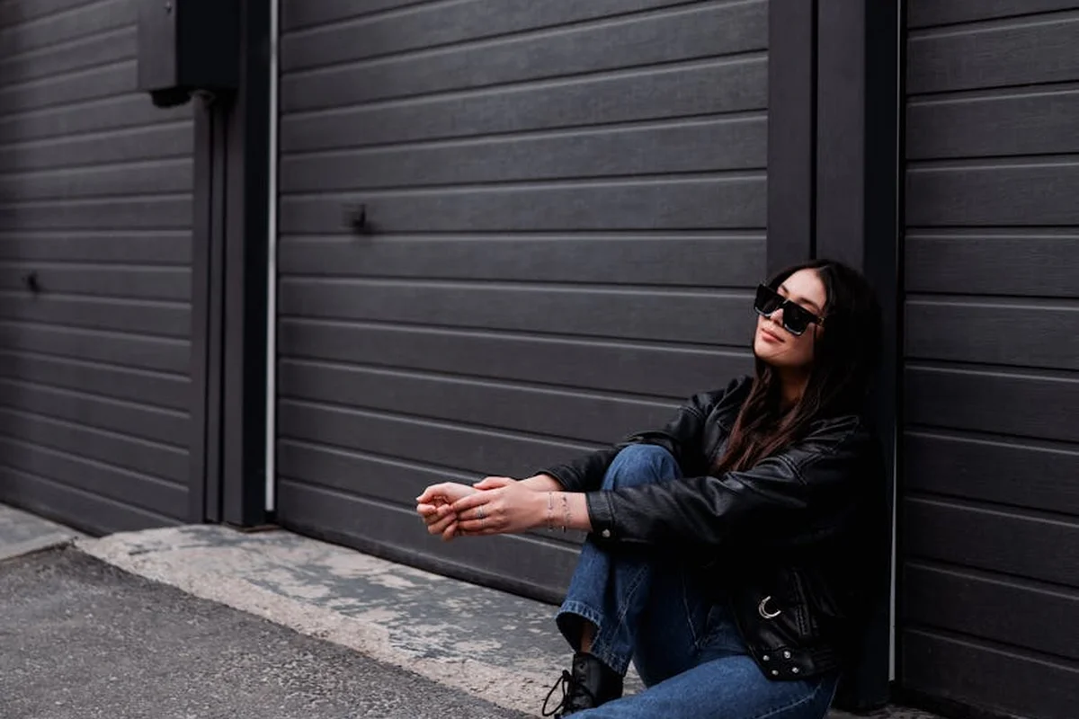 Person wearing a black jacket and sunglasses sits on the ground beside a closed dark garage door, preparing tools for garage door maintenance.