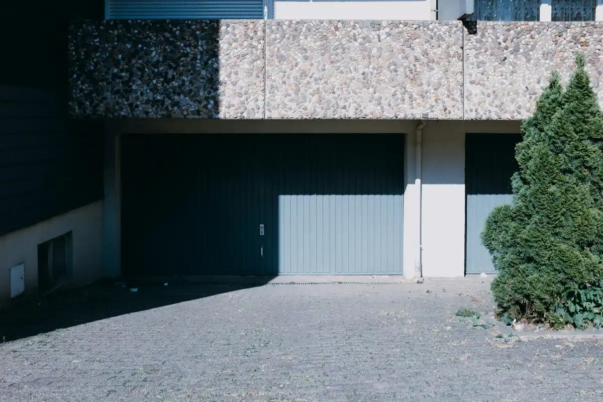 Residential garage with two blue doors, a stone facade, and a paved driveway with a tall evergreen shrub on the right.