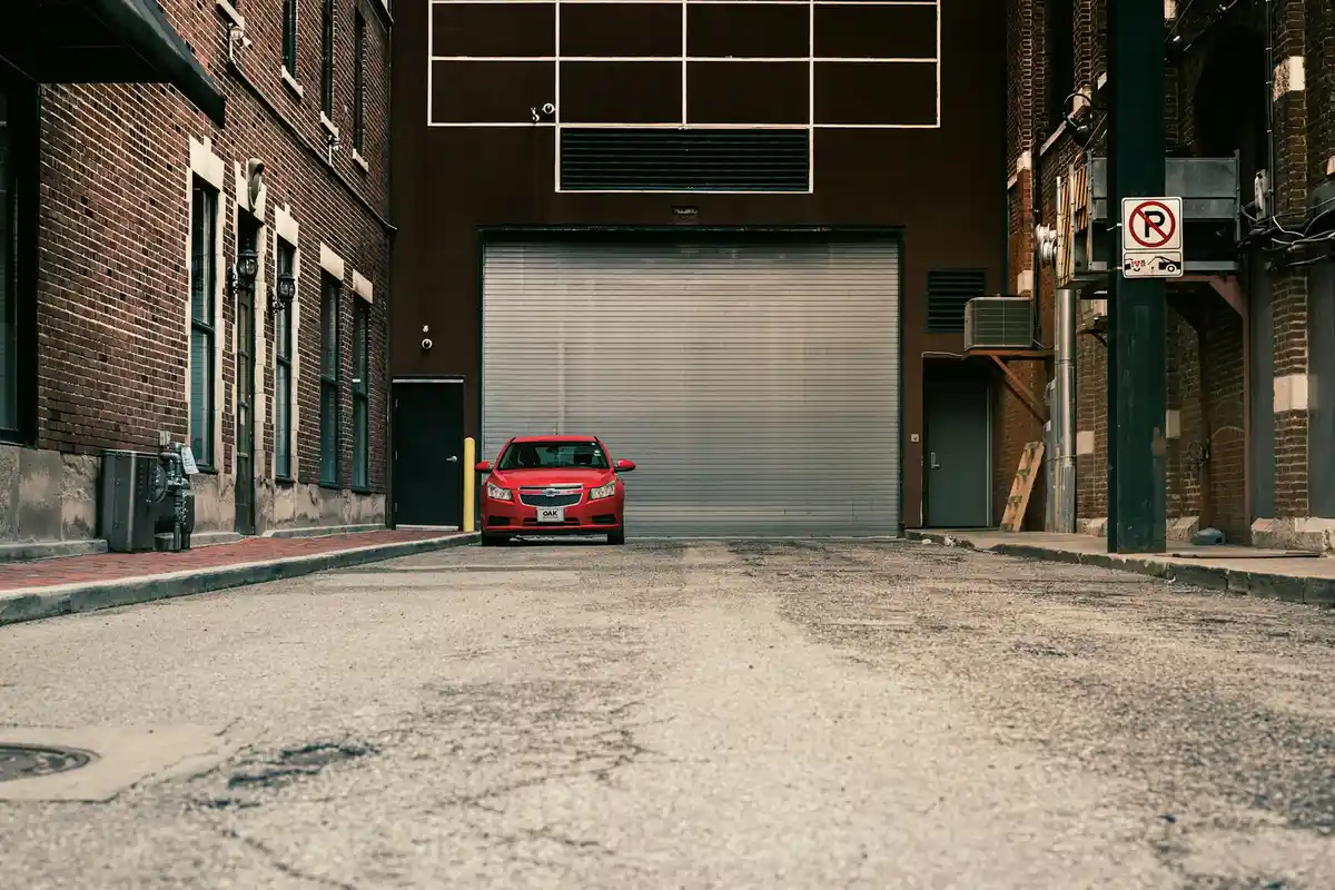 Urban alley scene with brick buildings and a large closed garage door at the end; a red car is parked near the center.