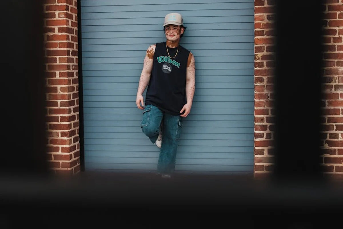 Tattooed person wearing a cap and sleeveless shirt leaning against a closed blue garage door between brick pillars.