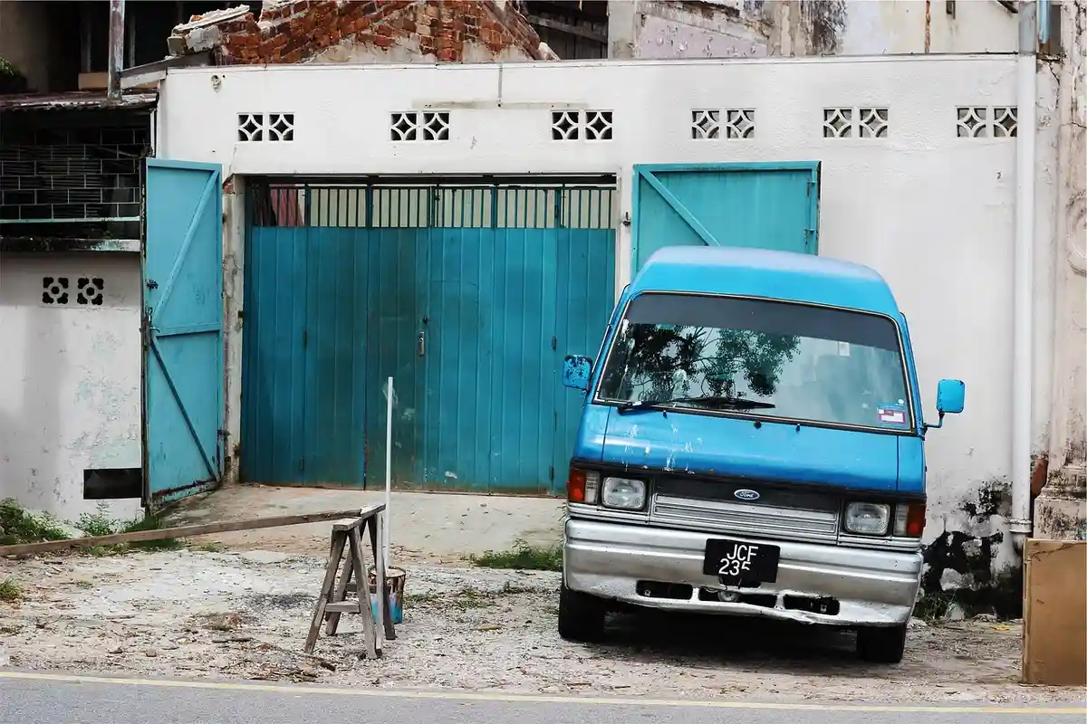 Outdoor scene with a blue van in front of a white building and blue garage doors; a small ladder sits on the ground, suggesting a workspace for installing a new garage door opener motor unit.