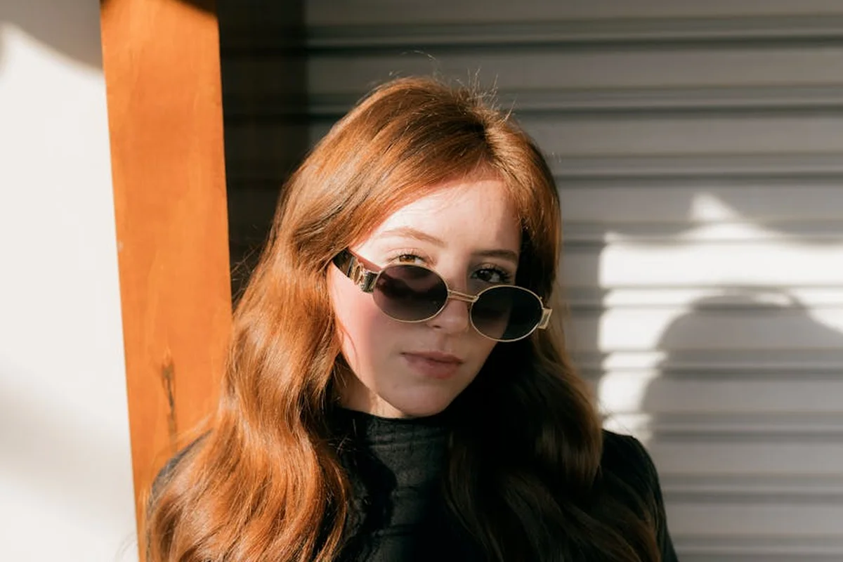 Person with long red hair and sunglasses standing beside a closed garage door.