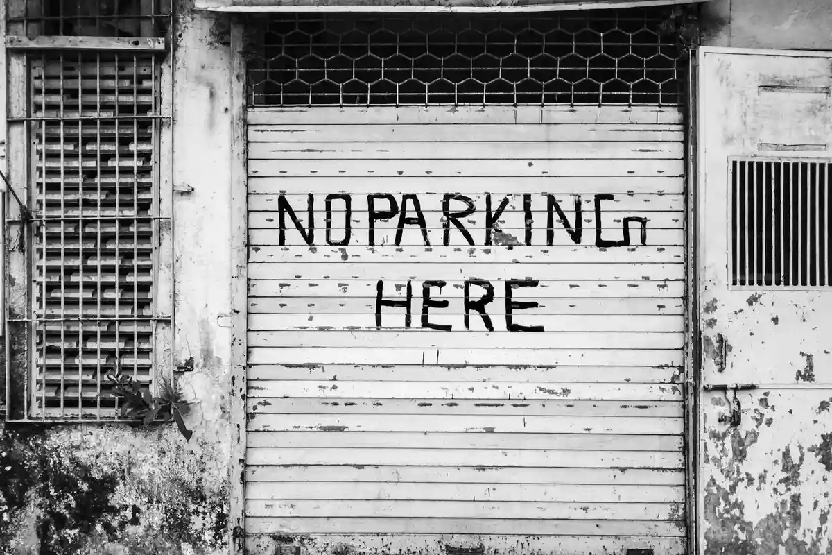 Black-and-white photo of a weathered garage door with peeling paint and rust, featuring a faded 'NO PARKING HERE' sign.