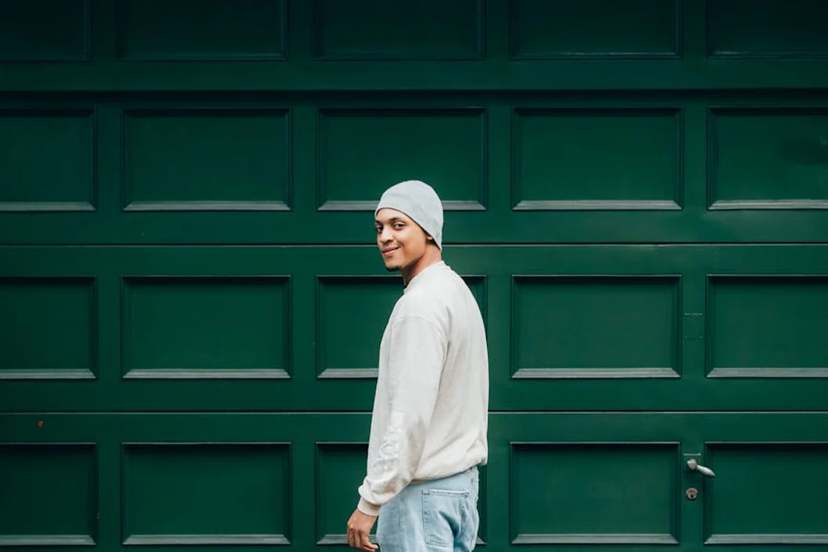 A person wearing a light-colored sweater and beanie stands in front of a green garage door, illustrating safety prep for removing a garage door from its tracks.