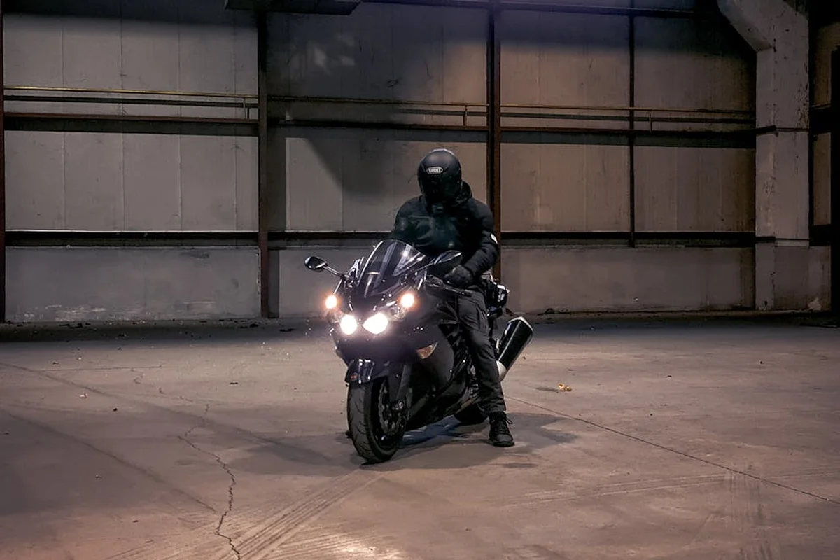A motorcyclist wearing a helmet sits on a sport bike inside a dimly lit, empty garage.