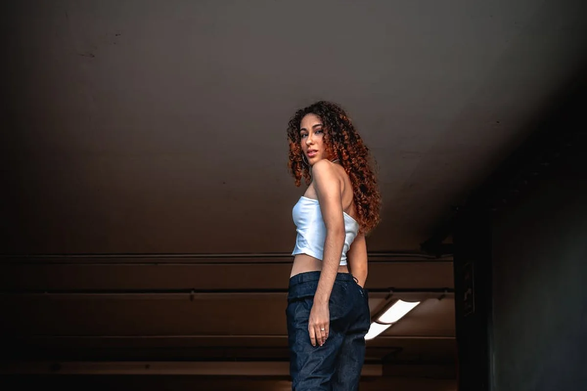 A person in a dimly lit garage wearing a white crop top and dark jeans stands near the doorway.