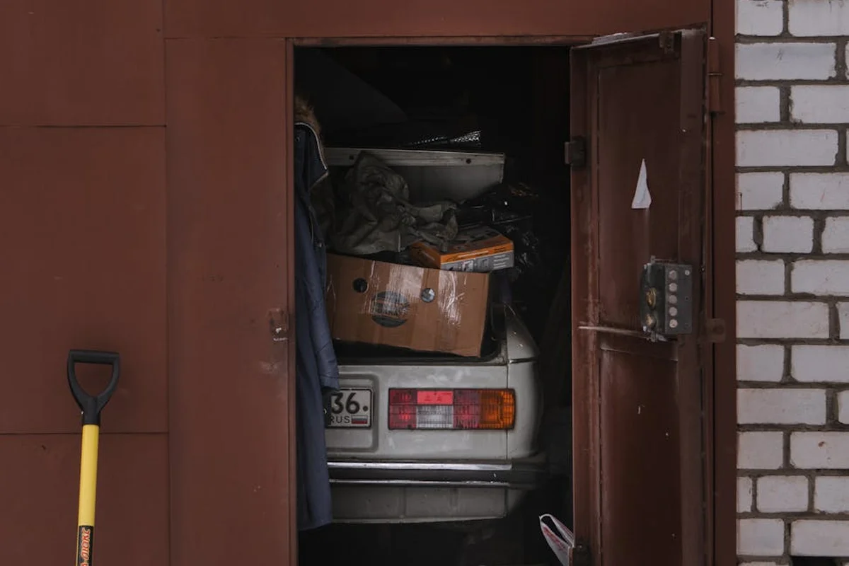 An open garage with a car parked inside, cluttered with boxes and tools, with a shovel leaning against the wall.