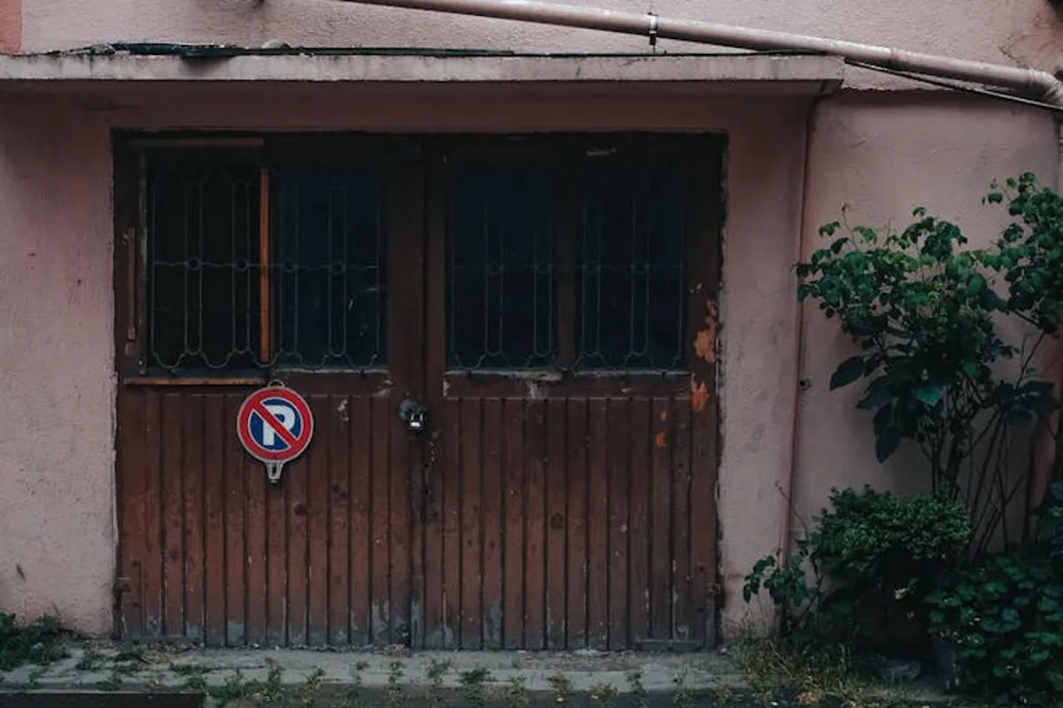 Weathered wooden garage doors with barred windows, a no-parking sign on the door, pink exterior walls, and greenery on the right.