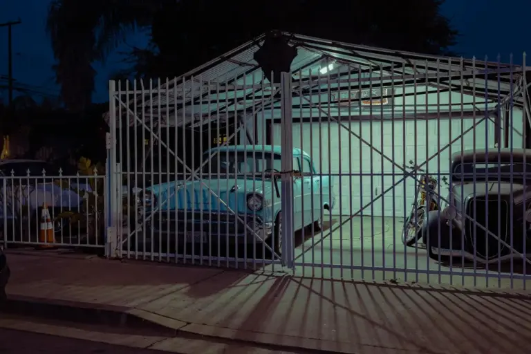 Nighttime gated driveway with a blue vintage car inside the garage and a motorcycle outside the gate.