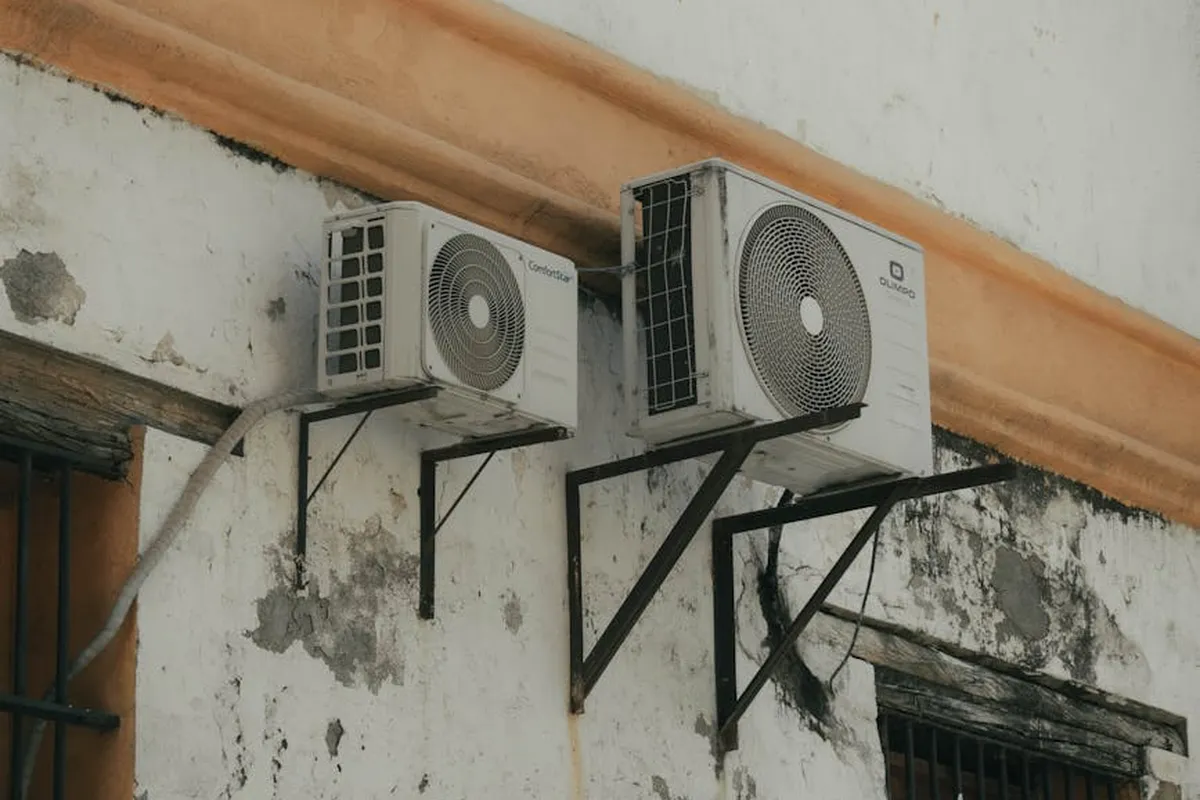 Two outdoor air conditioning condensers mounted on a weathered garage exterior wall with exposed pipes and mounting brackets.