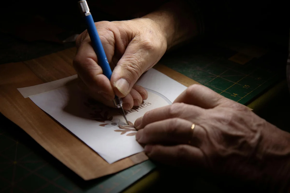 Close-up of hands tracing a leaf motif with a pencil on paper, preparing a stencil design on a work surface.