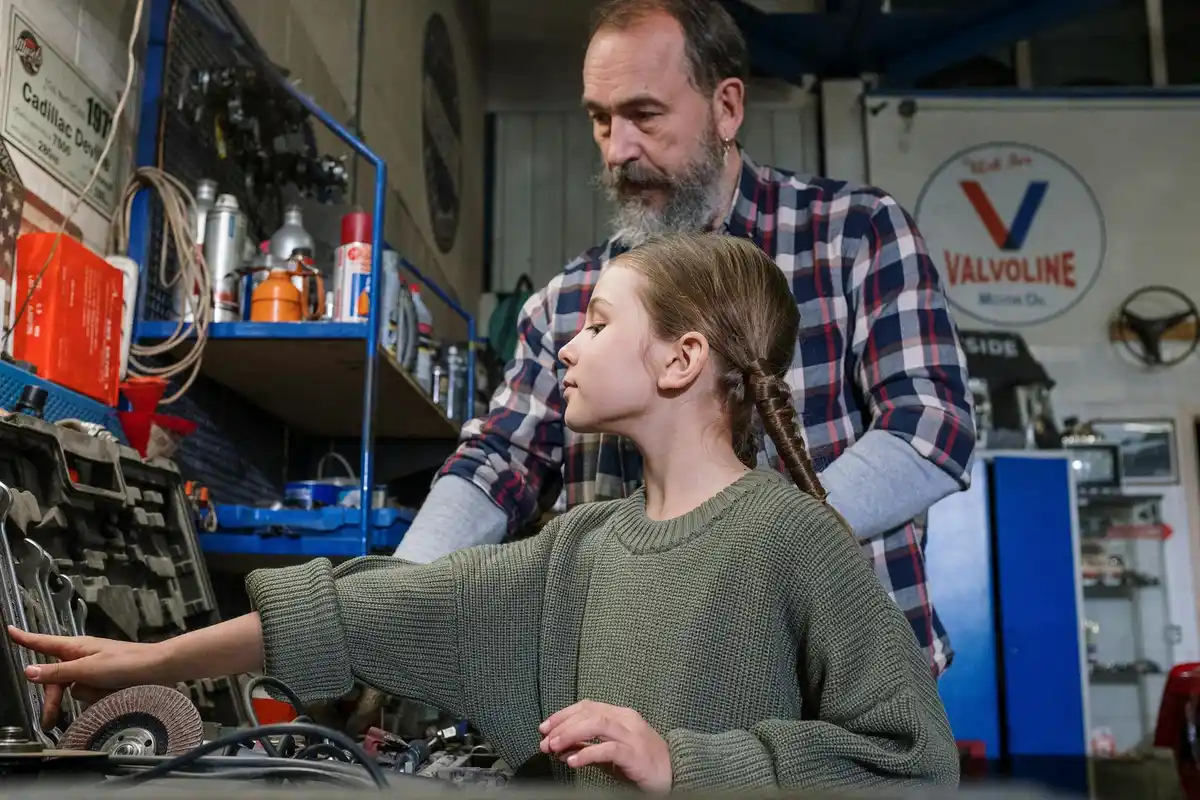 An adult and a young person work at a garage workstation, examining tools and equipment in a workshop setting.