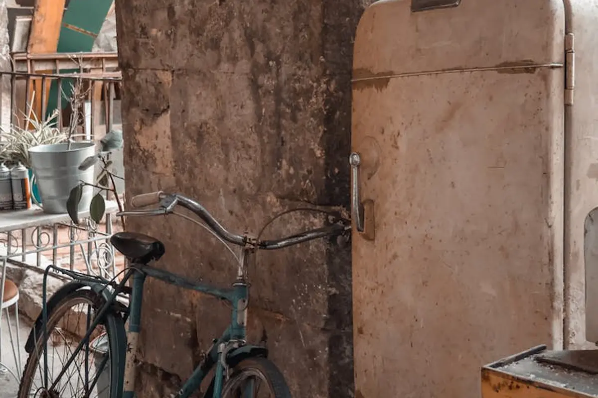 An old vintage refrigerator is placed in a dusty garage next to a bicycle and scattered tools.