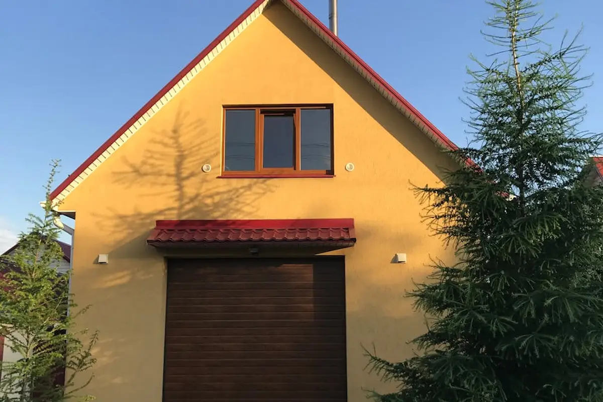 Front view of a yellow garage with a steep gable roof, red trim, and a large brown overhead door, with trees nearby.