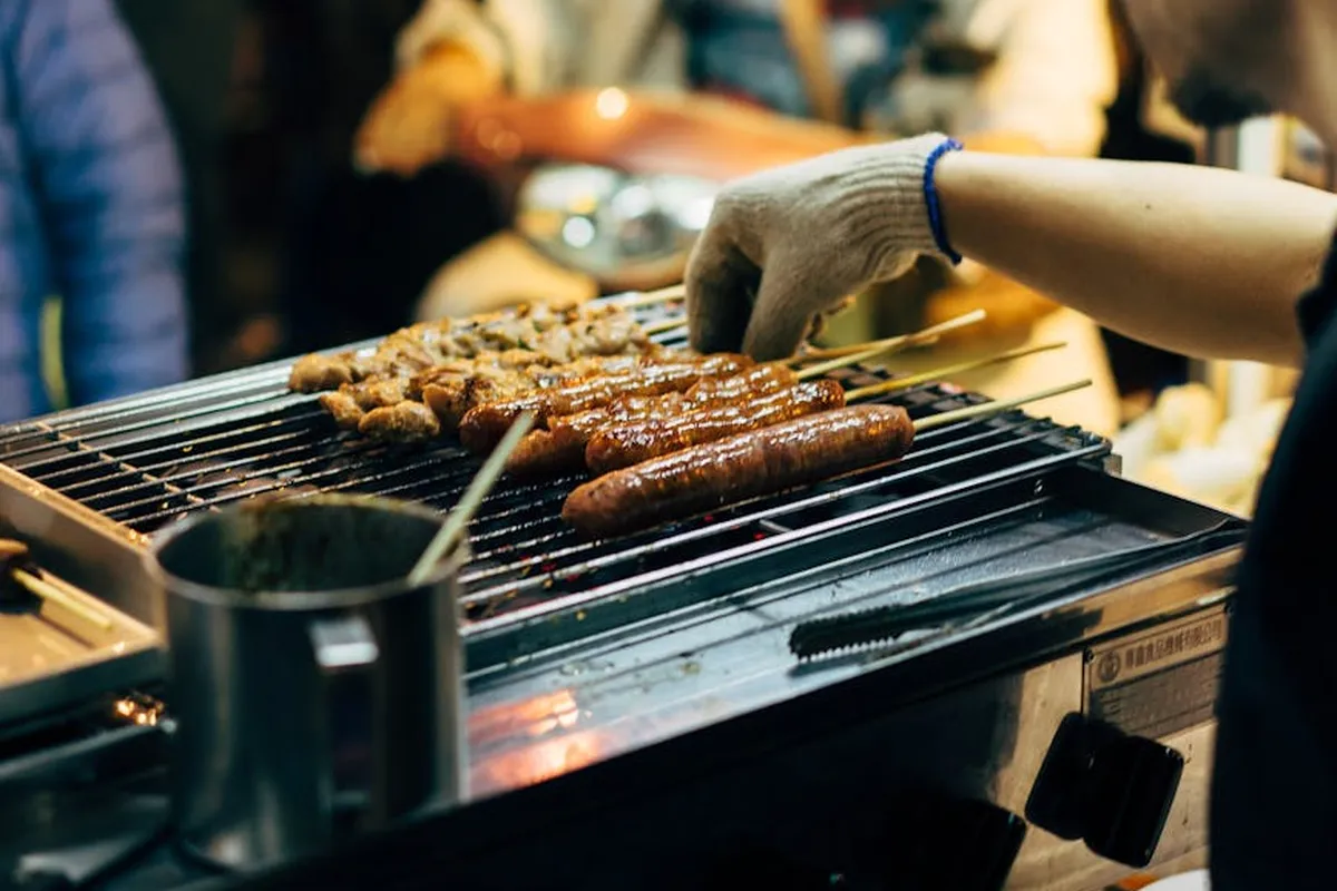 Grill with sausages and pieces of meat being cooked, a gloved hand tending the food in a garage-like indoor setting