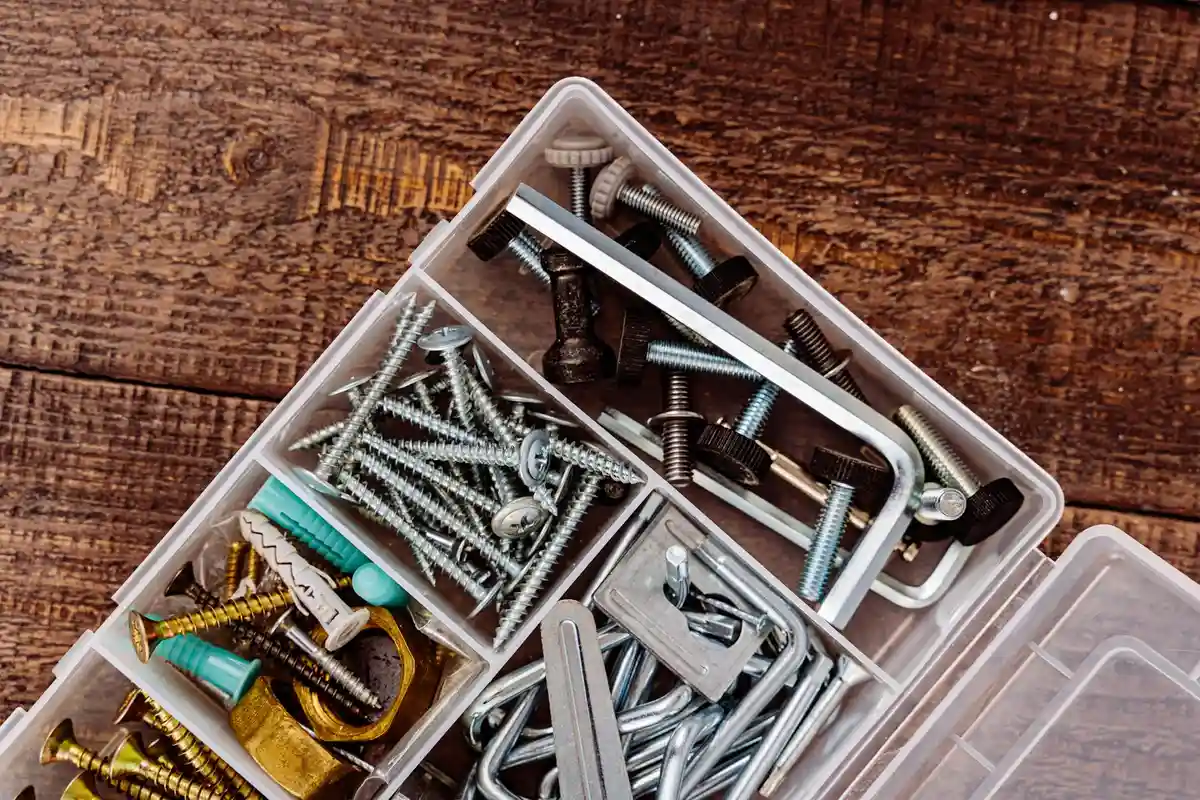 Clear plastic hardware organizer with multiple compartments filled with screws, bolts, nuts, and brackets on a wooden work surface