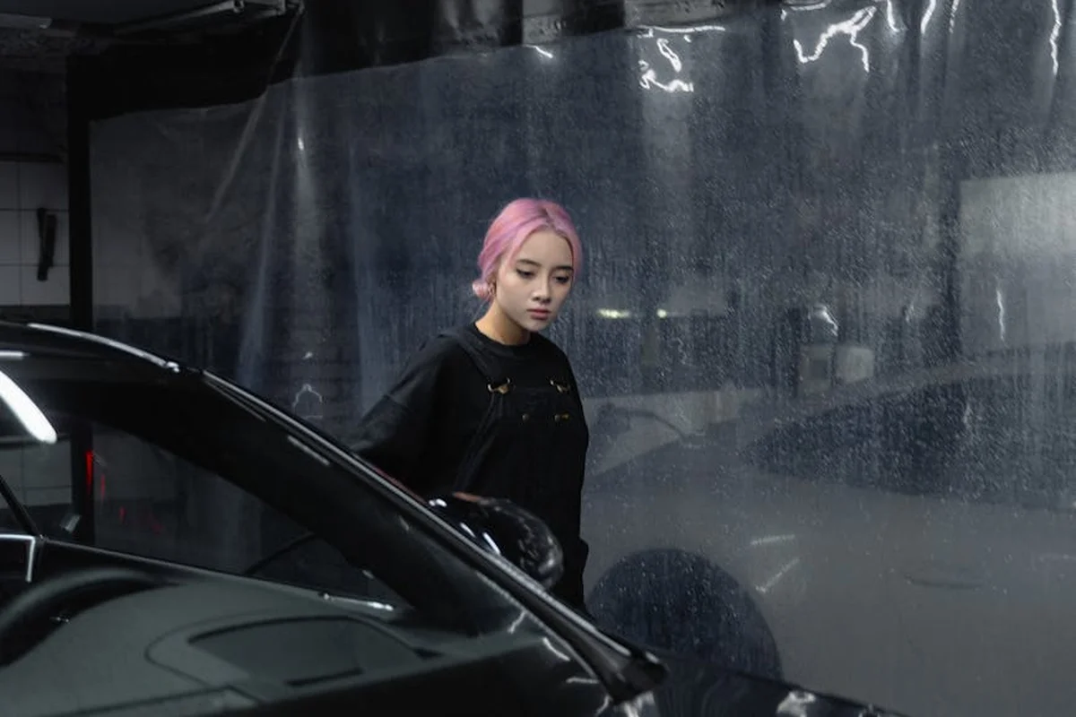 A person with pink hair stands near a car in a dim garage, examining tools for a heater installation.