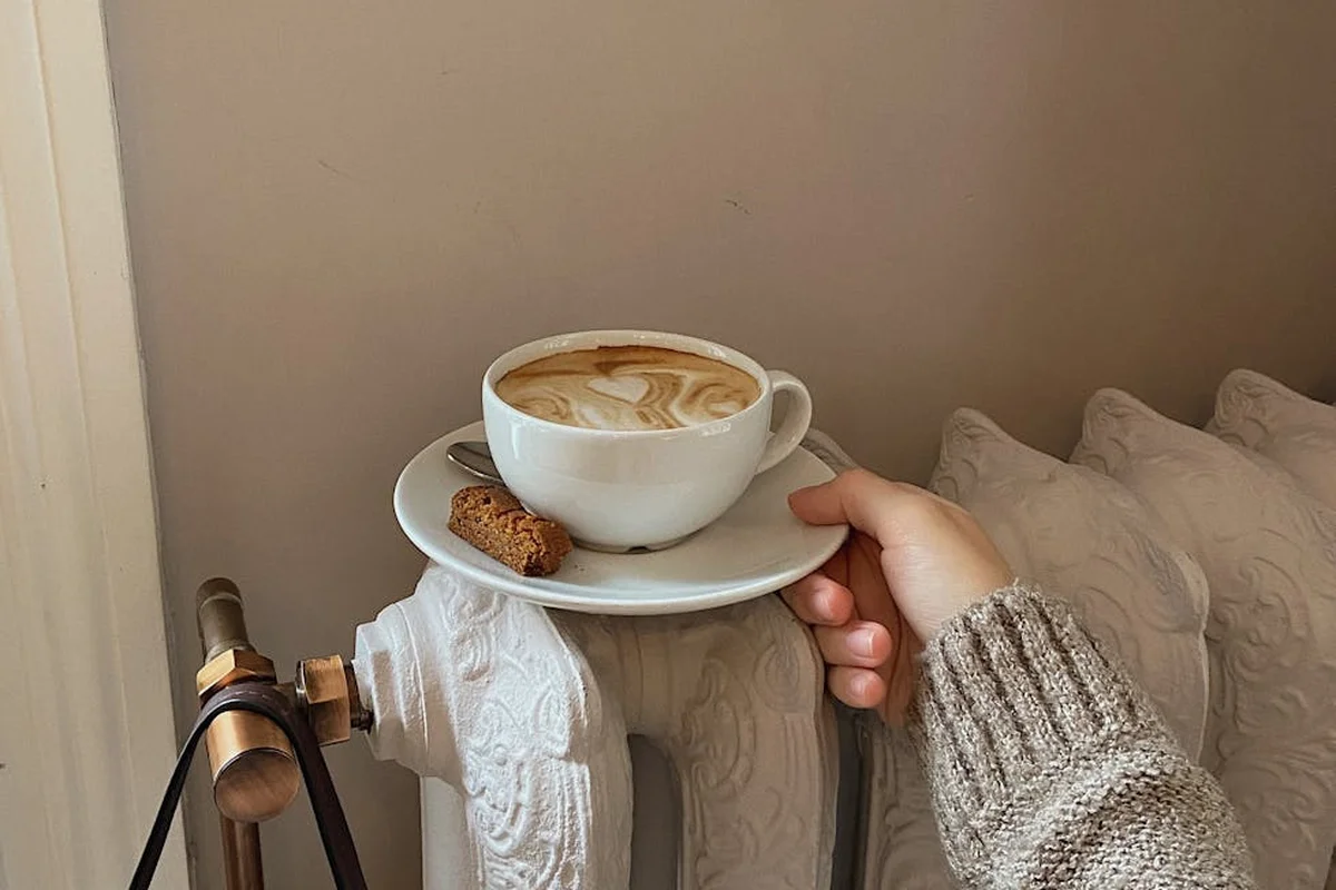 Close-up of a hand in a knit sleeve resting a cup of coffee on a saucer atop a radiator, illustrating a potential hazard around heating equipment.