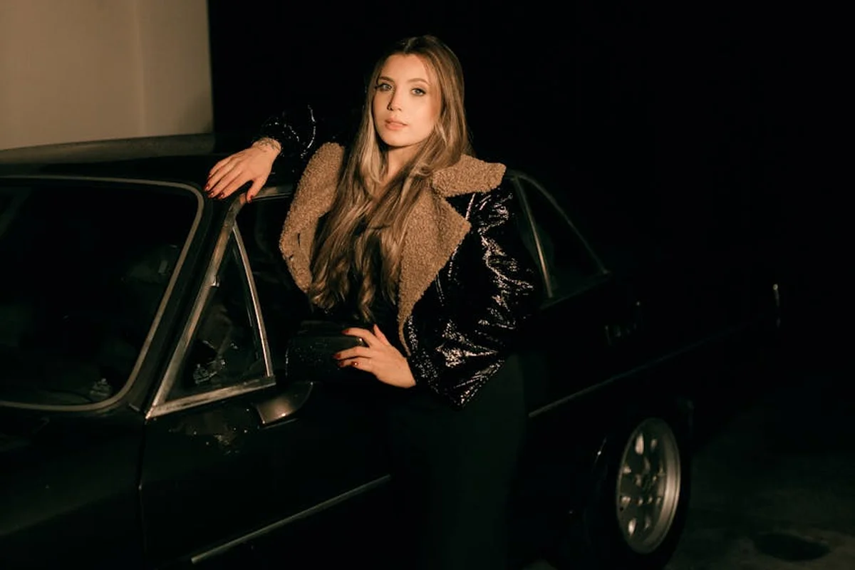 A woman with long hair, wearing a tan faux-fur-collared jacket and black outfit, stands beside a dark car in a dimly lit garage.