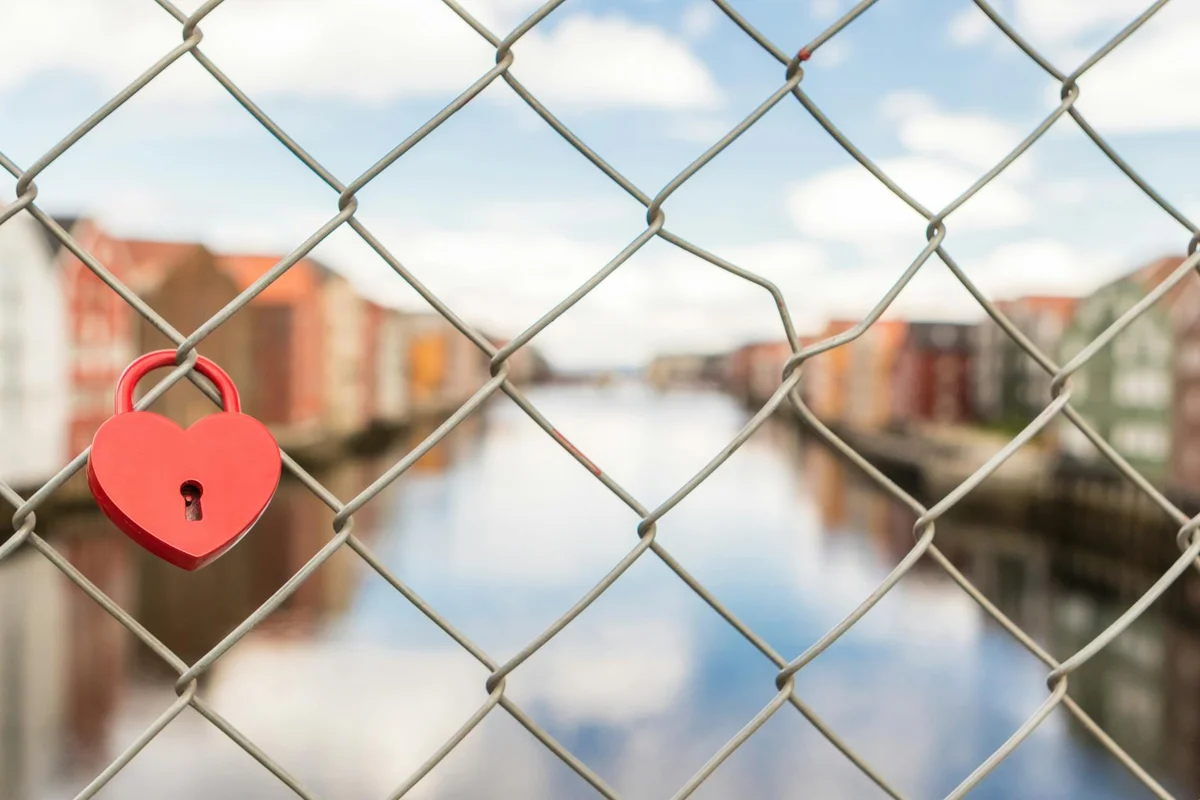 Red heart-shaped padlock hanging on a chain-link fence with a blurred urban background