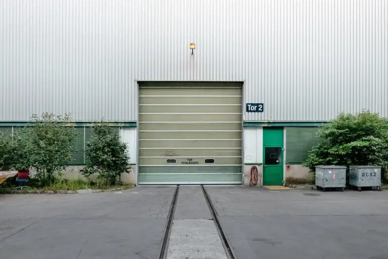 Industrial garage with a large metal roll-up door and a smaller green pedestrian door labeled Tor 2; concrete ground with rail tracks and nearby trees and dumpsters, illustrating potential gaps that mice could use to gain entry.