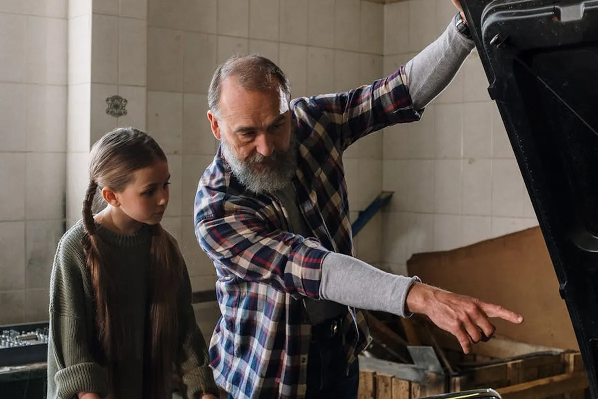 An adult man and a young girl stand in a garage with the hood of a car open, as the man points to something in the engine bay.