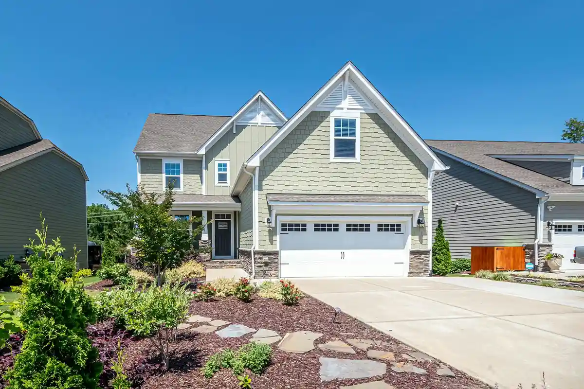 Two-story suburban house with a double garage, concrete driveway, and landscaped front yard under a clear blue sky.