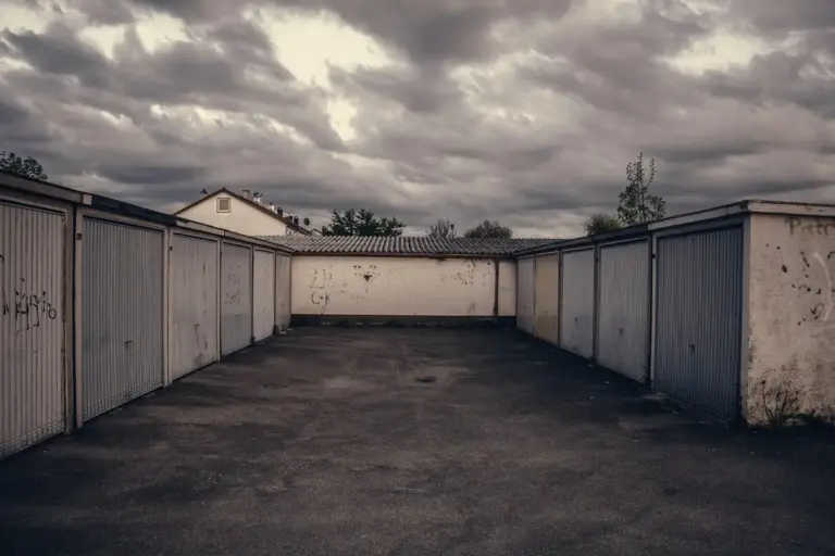 A long row of metal storage garages with closed doors, a paved driveway, and a cloudy sky.
