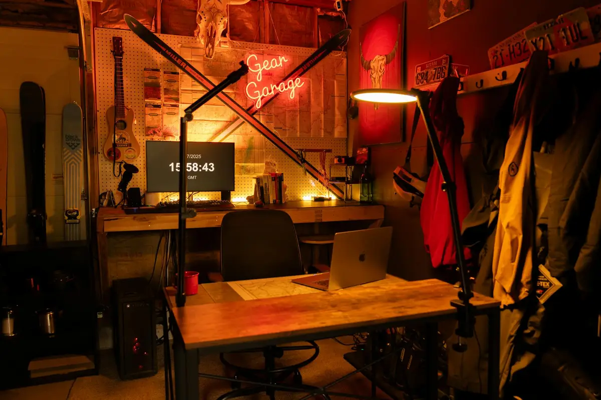 A small home garage recording space with a wooden desk, laptop, monitor, a microphone arm, and a neon 'Gear Garage' sign on the wall.