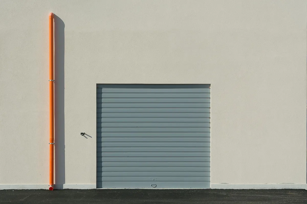 Exterior garage wall with a gray roll-up door and an orange vertical downspout against a beige facade.