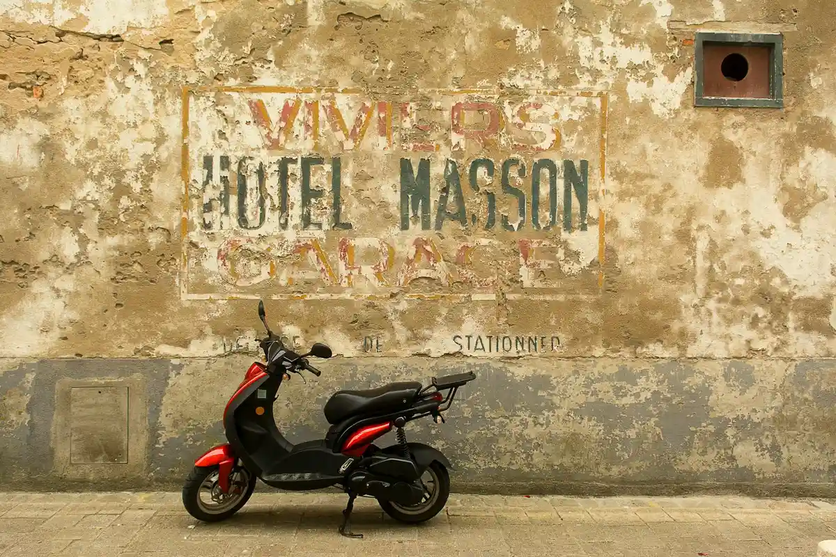 Weathered exterior wall with a faded sign and a red and black scooter parked on a sidewalk.