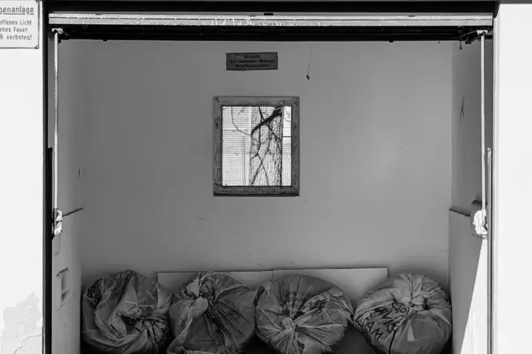 Small square window in the back wall of a garage opening to the interior, with bags stacked on the floor beneath it.