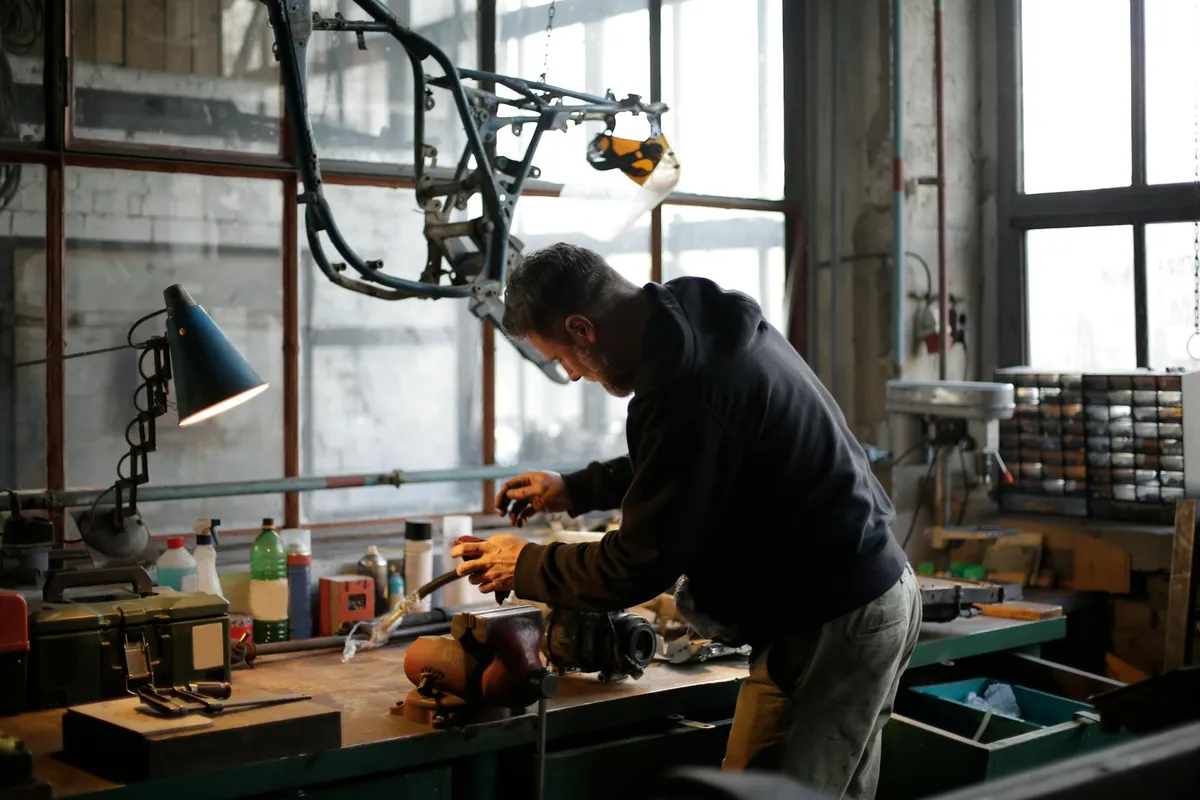 An individual in a dark hoodie leans over a cluttered workshop workbench, examining a component among various tools and materials.