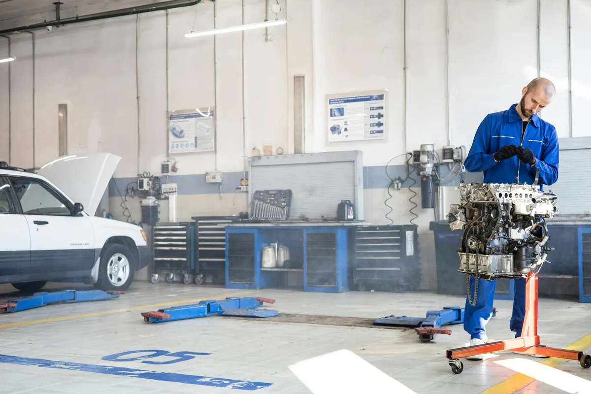 Technician in blue coveralls inspecting equipment in a busy garage workshop with a car on a lift and tools in the background.