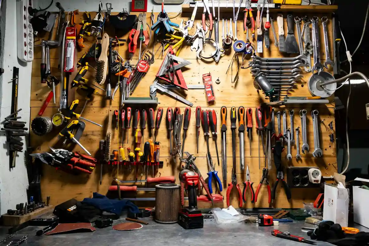 A busy garage workshop with a pegboard wall full of hand tools, screwdrivers, pliers, wrenches, and a cluttered workbench below.