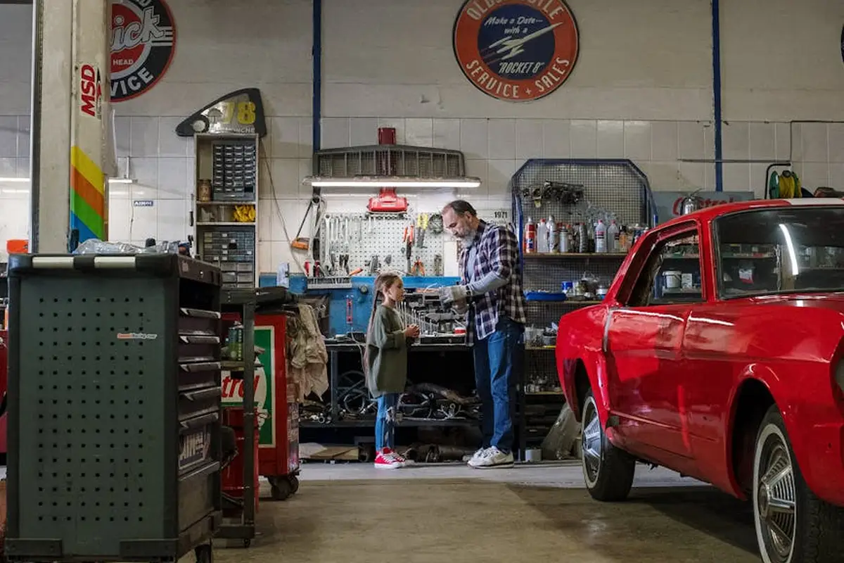 A mechanic assists a customer in a busy auto repair shop beside a red car, with organized tools and equipment in the background.