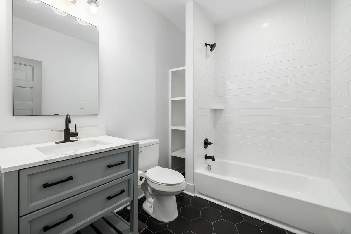 Modern compact bathroom in a garage renovation featuring a gray vanity with white countertop, a wall-mounted mirror, a white toilet, and a white tub/shower with dark fixtures.