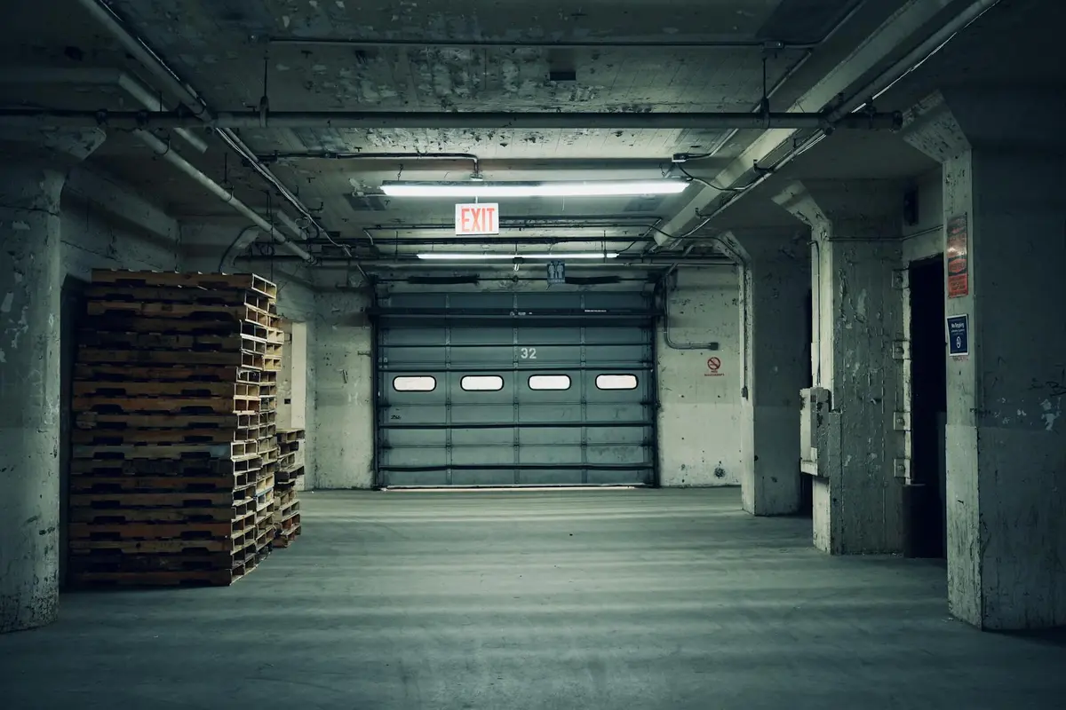 Industrial concrete garage interior with a closed sectional garage door, exposed ceiling pipes, and pallets stacked on the left.